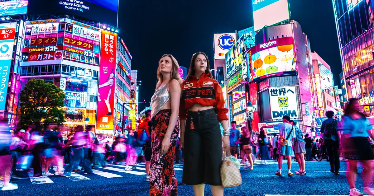 Two women posing at Shibuya Crossing at night with the neon screens behind them, on a Tokyo photoshoot
